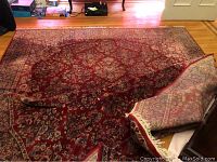 Full view of the cranberry tone area rug showing intricate floral traditional pattern with visible wear and some folding on one side.