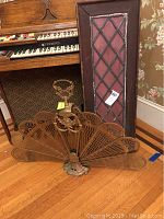 Brass fireplace fan in front of a vintage leaded glass window panel, displayed on a wood floor next to a musical instrument.