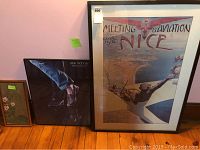 Photo showing three framed pieces of wall art leaning against a wall on a wooden floor: vintage 'Meeting Aviation Nice' print, 'Blue Monday' poster by Peter D. Esposito, and a small Asian floral art print.