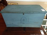 Front view of the blue painted wooden trunk showing surface wear and faint emblem on front.