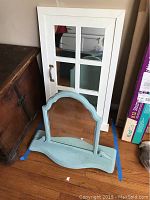 Two mirrors shown together on a hardwood floor: a white framed cabinet door mirror with panes and handle leaning against the wall, and a blue painted vanity top mirror with an arched top on the floor.