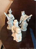 Three porcelain Asian themed statues on dark wood surface: woman with lute, man with lute, two children statue