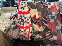 Six quilt tops folded and draped over furniture showing variety of patterns including star blocks, plaids, calico and stripes