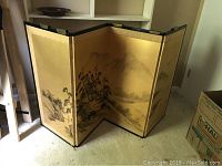 Photo showing three-panel vintage oriental folding screen with gold silk-like finish and rural landscape artwork, positioned partially folded in a room.