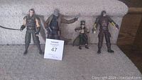 Four Arkham video game character action figures arranged on carpet in front of a carpeted step. Characters from left to right are Ra's al Ghul, Solomon Grundy, Mad Hatter, and Deathstroke.