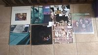 Photo showing eight vinyl record albums laid out on a tiled floor, visible covers with varied artwork including photos and illustrations.