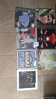 Photo showing seven vinyl records laid out on the floor, each with visible cover art, colors, and some text. Records include various artists and styles.