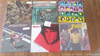 Photo showing six vinyl record albums with varied classic rock artists and colorful covers on a wooden floor.