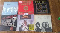 Photo showing six vinyl records in jackets laid out on a wooden surface. Includes albums by The Beatles, Jefferson Starship, The Fools, The Doors, and others.