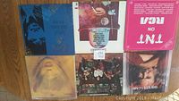 Photo showing six vinyl records arranged on a wooden floor with varied album covers including blue, dark, bright pink, sepia tones, and group portrait designs.
