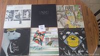 Photo shows six vinyl records on a wooden surface, featuring covers for Genesis, Eagles, The Fugs, The Good Rats, a record with an Asian mask themed cover, and a Bluebird Blues record with yellow label.