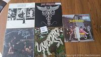 Photo showing five vinyl records with varied jacket artwork and band photos on wooden floor, condition of jackets visibly varied.