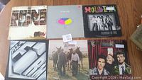 Top view of six vinyl records with visible album covers by Nine, Vixen, Ratt, Fastway, The Doobie Brothers, and Oingo Boingo, placed on a wood floor.