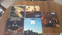 Five vinyl records displayed on wooden table. Album covers visible include The Cars, Eagles, Jethro Tull, and Moody Blues.