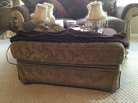 Brown fabric ottoman with wheels, shows wear. Positioned in front of a sofa with two fringed lamps behind it.