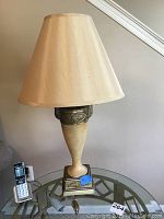 Lamp on glass table next to cordless phone, showing ceramic base with metal adornment and cream silk-like lampshade.