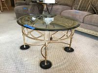 Full view of round glass top coffee table on carpet, showing metal base with geometric design and black round feet.