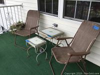 View of two folding chairs and two vintage metal side tables on a balcony with green outdoor carpet, showing wear and rust on tables and fabric wear on chairs.