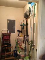 Room with wood and metal folding step ladders, multiple brooms, dustpans, mop with bucket, and shovel leaning against the wall