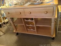 Front view of light wood server showing one drawer, three divider sections including built in wine rack, and four caster wheels.