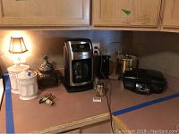 Countertop view showing Farberware coffee maker in stainless steel and black plastic, electric grill, metal and plastic ice bucket, salt and pepper shakers, glass canister with metal lid, and lamp.