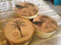 Three round decorative hat boxes; two with cherub lids, one with palm tree and vintage postcard print. Boxes vary in size.