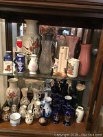 Five shelves of various porcelain and glass collectible vases and figurines including some decorated with floral patterns and a distinctive nutcracker-style figurine.