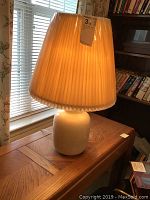 Front view of the ceramic lamp on a wooden table illuminated by daylight, showing the classic shape and pleated shade.