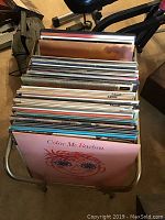 Large collection of vinyl LP records stored in a metal or plastic record holder rack showing numerous LP records with colorful sleeves stacked vertically, visible wear.