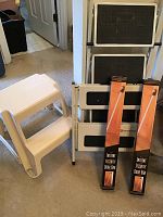 Photo shows white plastic two-step stool, two foldable metal step ladders with black treads, and two instant security door bars in packaging leaning against a wall.
