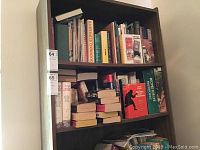 Photo showing top two shelves of a bookcase filled with assorted books including 'Illustrated Cookbook' and others, stacked vertically and horizontally.