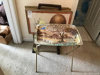 Photo showing one TV tray with a painted rustic cabin and trees scene, set on a metal folding frame. In the background several additional trays and frames are visible.