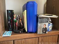 Office supplies organized on wooden cabinet including Suze Orman Financial Freedom book and kit, blue file box, pens, pencils, markers, sticky notes, and spiral notebook in desktop organizer.