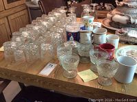 Top view of approximately 65 pieces of glassware and cups including tumblers, mugs, and stemmed glasses displayed on a wooden table.