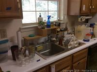 View of kitchen sink and counter area with various plastic cups, glasses, trays, and cleaning supplies visible on counter and window ledge.