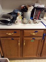 Photo showing a George Foreman grill, multiple ceramic mugs with floral patterns, tea cups and saucers, kitchen utensils in a container, and a portion of oak kitchen cabinetry with drawers.
