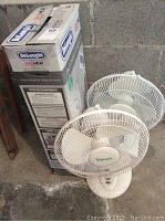 Two white oscillating pedestal fans next to the boxed heater, one fan showing brand 'Emerson' on center grille, fan controls visible on base.