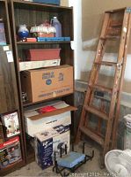 Photo showing laminate book shelf with cardboard boxes, plastic containers, wooden ladder leaning against wall, and various items on shelves and floor.