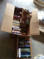 Open cardboard boxes filled with paperback and hardcover novels stacked vertically and horizontally showing spines and covers of various titles and authors.