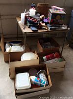 Plastic containers and miscellaneous items on top of and around folding card table with boxes on floor