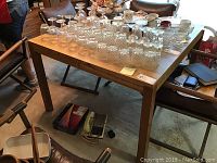 Full view of rectangular wooden dining table with various glassware on top, surrounded by chairs. The table top is a light to medium brown wood finish.