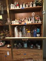 Wooden cabinet shelf displaying multiple porcelain figurines including flower themed ceramics, a stemware set, and an ice bucket.