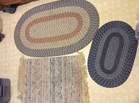 Three small rugs: two oval braided rugs in earth tones and one rectangular rag rug with fringes.