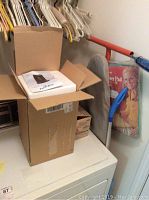 Open box with Air Innovations humidifier instruction manual on top of styrofoam packaging inside the box placed on a washer or dryer.