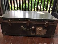 Front view of dark green military foot locker with reinforced metal corners and two metal latches. Shows surface wear, torn label remnants on front corner, and central lock.