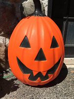 Front view of large orange blow mold jack-o-lantern with black facial features, showing triangular eyes, nose and jagged smiling mouth.