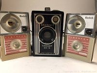 Front view of three cameras: two Kodak Hawkeye Flashfun cameras with flash attachments on left and right, Kodak Brownie Target Six-16 box camera in center