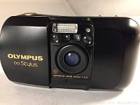 Front view of Olympus Stylus camera showing lens, built-in flash, and branding.