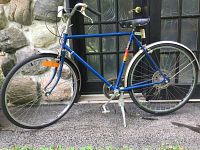 Side profile of the blue Supercycle bicycle showing the frame, wheels, crank, chain, pedals, and saddle in outdoor setting.