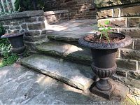 Two cast iron urns situated on stone steps beside a stone wall, both showing signs of age and wear.